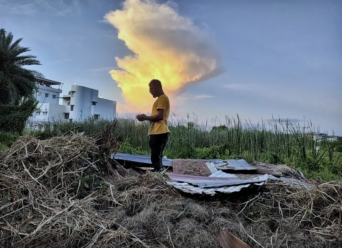 A man in a yellow shirt standing on a heap of dry grass as a tall cloud glows gold behind him